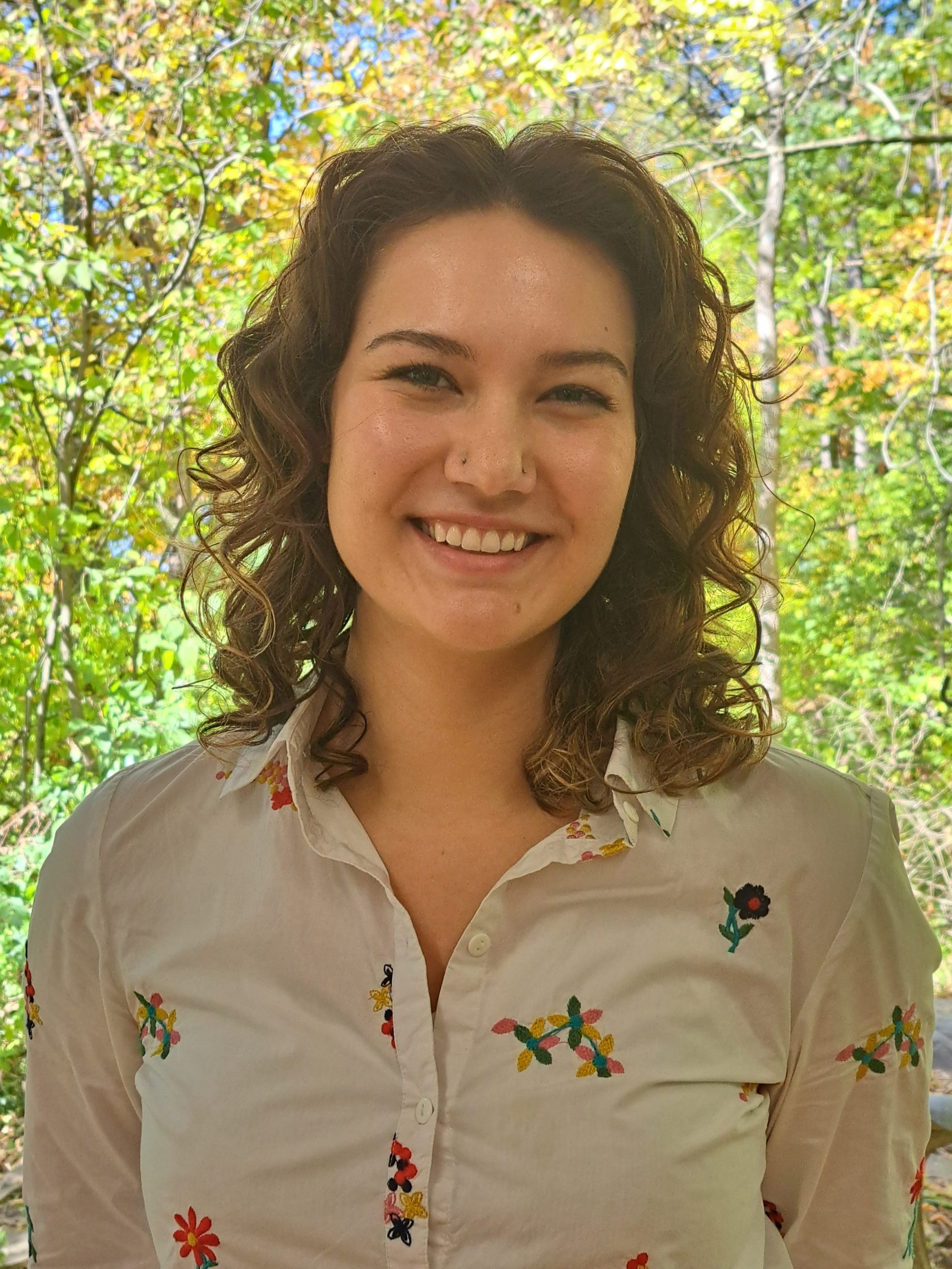 Anita Jackson smiling in a white shirt. Trees and shrubs behind her. 