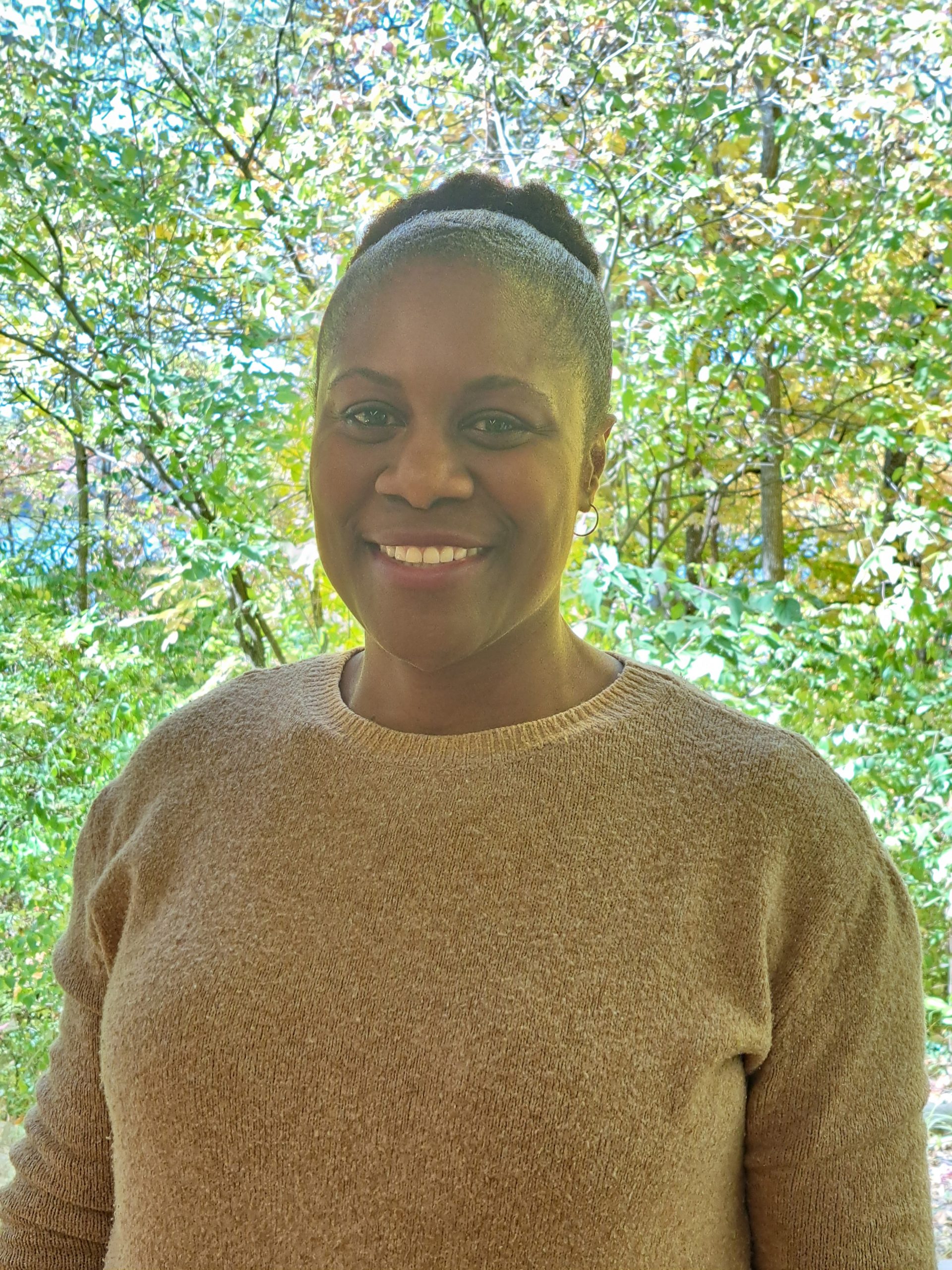 Anita Jackson smiling in a white shirt. Trees and shrubs behind her. 