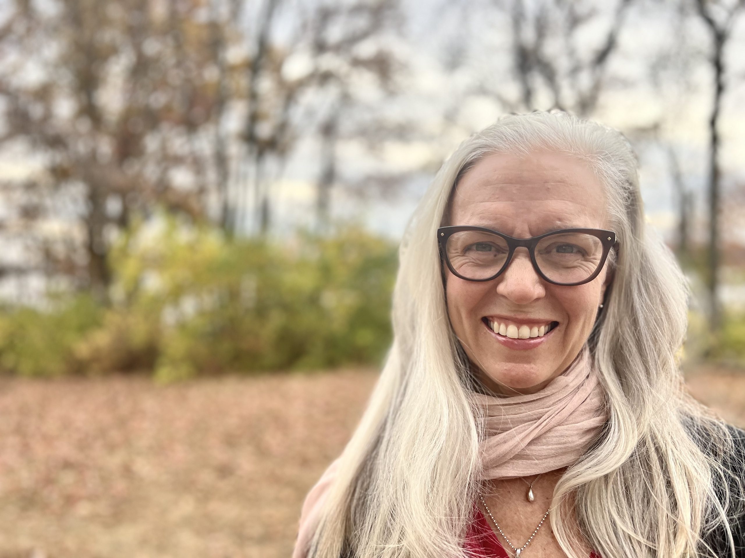 Anita Jackson smiling in a white shirt. Trees and shrubs behind her. 